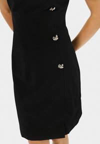 Robe noire arrivant au genou avec trois fermoirs décoratifs en argent en forme de papillon le long du côté droit, portée par une personne les bras croisés derrière le dos.