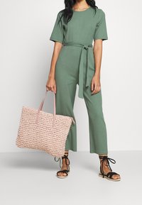 Groene geribbelde jumpsuit met een ceintuur in de taille, gecombineerd met een grote roze geweven tote bag en zwarte sandaaltjes met bandjes. Eenvoudig en moderne uitstraling.