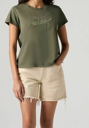 Vrouw draagt een olijfgroene Levi's 73 T-shirt en beige denim shorts met rafelige zoom, staand met één hand in de zak.
