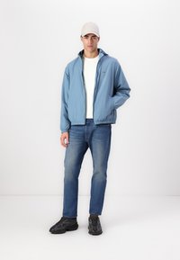 Lyseblå lett jakt med hette, elastiske ermer og midje. Brukt over en hvit t-skjorte, kombinert med blå jeans og svarte joggesko.