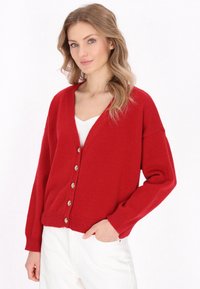Červený pletený cardigan s výstřihem do V, předními knoflíky a žebrovanými manžetami. Měkká textura a uvolněný střih, kombinovaný s bílým topem a světlými kalhotami.