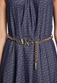 Femme portant une robe bleue à motifs avec une ceinture chaîne dorée à boucle « MK » et une étiquette suspendue gravée « Michael Kors ».