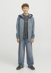 Hallhalli, kerge kapuutsiga jakk, millel on sile tekstuur, kantud mustrilise tumeda särgi ja vabalt istuvate siniste teksade peal. Välimuse lõpetavad pruunid kingad.