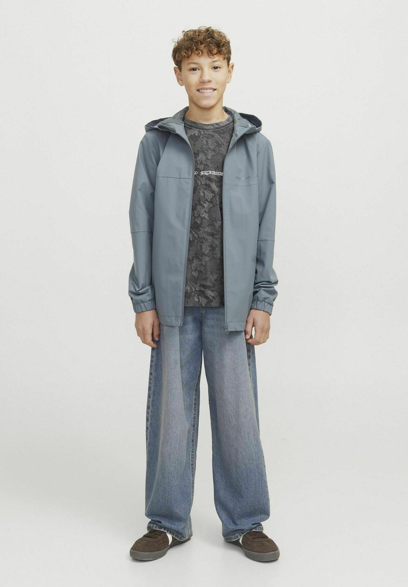 Hallhalli, kerge kapuutsiga jakk, millel on sile tekstuur, kantud mustrilise tumeda särgi ja vabalt istuvate siniste teksade peal. Välimuse lõpetavad pruunid kingad.