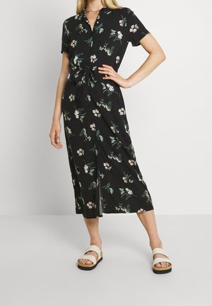 Kobieta w czarnej, kwiecistej sukience midi z krótkimi rękawami i białych sandałach, stojąca z jedną ręką na biodrze.