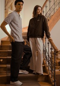 Dva modely na schodech: jeden v šedém polo tričku a černých kalhotách, druhý v hnědé bomber jacket a světlých šedých rozšířených kalhotách.