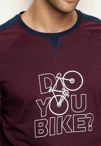 Camisa de manga larga color burdeos con un gráfico de bicicleta blanca y el texto "¿HACES BICICLETA?" en contorno de líneas discontinuas. Acentos en el hombro en color azul marino.