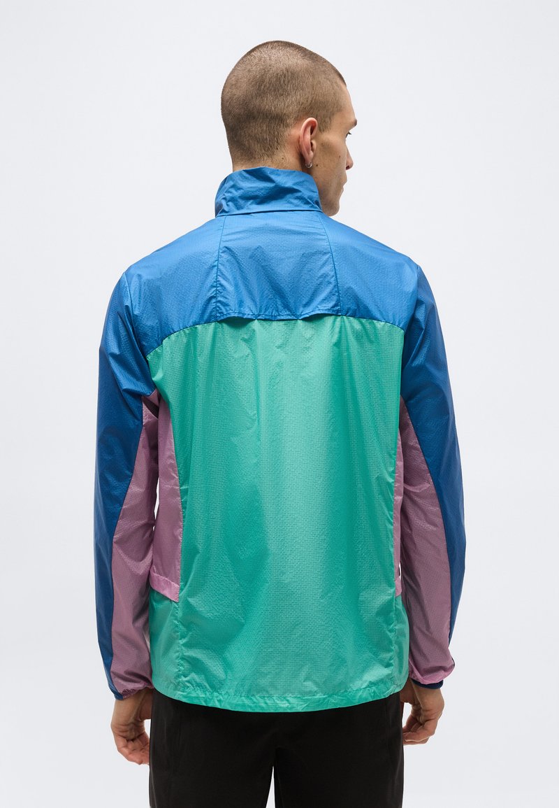 Mann trägt eine Windjacke mit Farbfeldern in Blau, Türkis und Lila, ist mit dem Rücken zu einem weißen, schlichten Hintergrund gewandt.