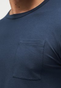 Κοντινό πλάνο ενός ατόμου που φοράει μπλε σκούρο μπλουζάκι με τσέπη στο στήθος και ορατή ραφή σε λείο ύφασμα.