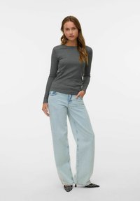 Jeune femme debout, une main dans la poche, portant un haut à manches longues gris foncé, un jean large bleu clair et des talons noirs à motifs.