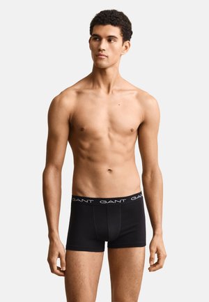 Młody mężczyzna stoi na białym tle, nosząc czarne bokserki marki GANT, patrzy na bok w zrelaksowanej pozie.