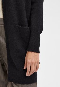 Cardigan en tricot noir avec une texture douce, comportant une poche latérale et des poignets côtelés. Le tissu semble chaud et confortable.
