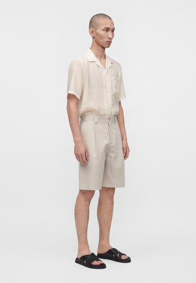 Homme portant une chemise beige clair à manches courtes, un short assorti et des sandales noires à bouts ouverts, debout devant un fond blanc uni.