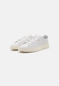 Vita lädersneakers med en slät textur, rund tå, platt beige sula och subtil branding på sidan. Snören är vita, med en kontrasterande hälflik i mörkt.