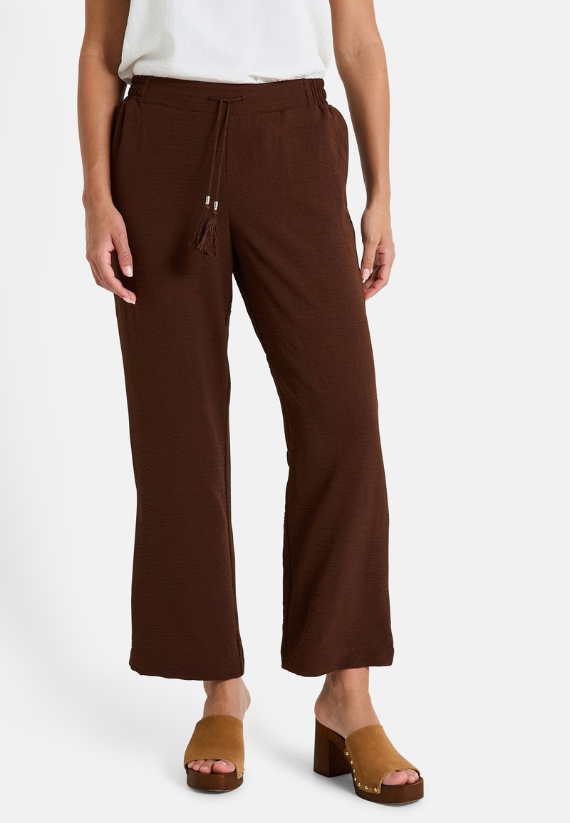 Une personne se tient debout, portant un pantalon large marron avec une taille élastique, associé à des sandales à talons carrés beiges. L'arrière-plan est blanc uni.