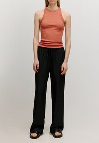 Ein rustfarbener, figurbetonter Tank-Top, kombiniert mit lockeren schwarzen Hosen. Das Tank-Top hat einen hohen Halsausschnitt und ein ärmelloses Design; die Hosen haben einen Kordelzug am Bund.