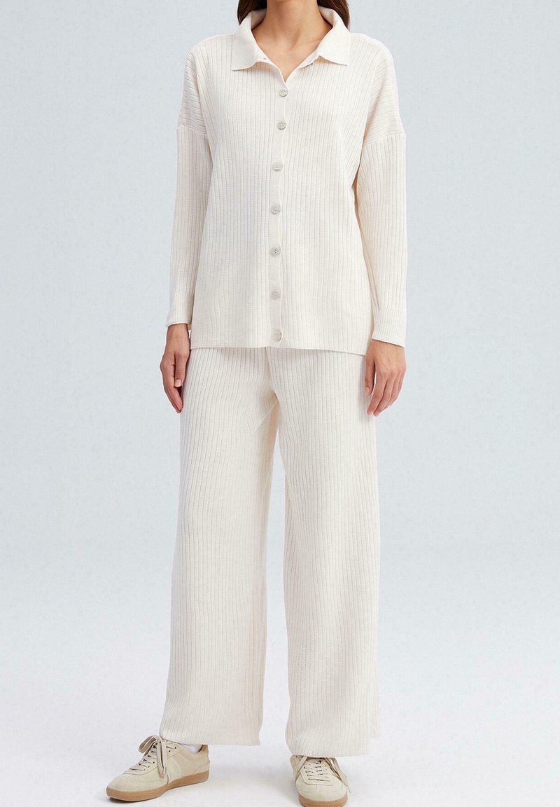 Femme portant un haut crème à côtes boutonné à manches longues et un pantalon large assorti avec des baskets beiges, debout sur un fond uni.