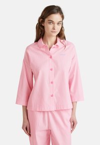 Camisa a rayas rosa y blanca con cuello, botón en la parte delantera y mangas de tres cuartos. Presenta una tela ligera y un sutil detalle de logo.