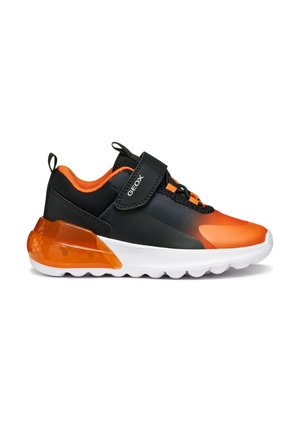 Chaussure de sport noire et orange avec une tige lisse, des accents texturés, une sangle en Velcro et un talon translucide orange. Semelle blanche avec des crampons.