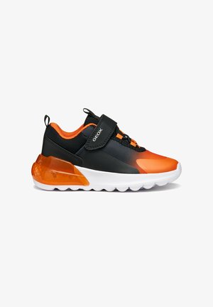 Chaussure de sport noire et orange avec une tige lisse, des accents texturés, une sangle en Velcro et un talon translucide orange. Semelle blanche avec des crampons.