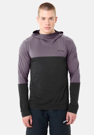 Giovane uomo con felpa con cappuccio bicolore viola e grigio scuro, in piedi contro uno sfondo chiaro uniforme, che guarda avanti.