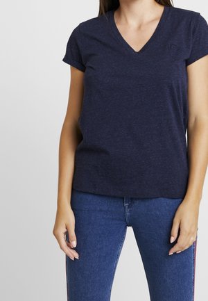 Kobieta ubrana w granatowy T-shirt z krótkim rękawem i dekoltem w serek oraz niebieskie dżinsy, stoi z ramionami swobodnie opuszczonymi wzdłuż ciała.