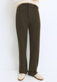 Pantaloni su misura verde oliva, dal taglio dritto e con pieghe frontali, caratterizzati da una texture liscia e dettagli di design sottili. Abbinati a scarpe chiare.