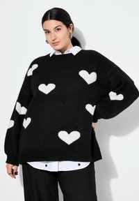 Schwarzer Pullover mit weißen Herzmustern, weiche Stricktextur, übergroßer Schnitt, lange Ärmel, getragen über einem weißen Hemd mit Herzakzenten.
