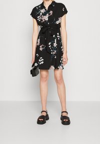 Schwarzes Blumenkleid mit kurzen Ärmeln, Kragen, Taillengürtel und ausgestelltem Rock. Kombiniert mit klobigen schwarzen Sandalen und einer Mini-Handtasche.