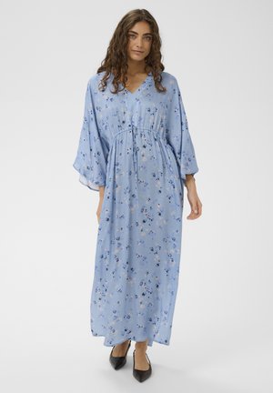 Vrouw met krullend haar, gekleed in een lichtblauwe, bloemrijke maxi-jurk met lange mouwen en zwarte pumps met puntige neus, staand tegen een effen achtergrond.