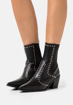 Bottines noires en cuir avec des pointes acérées, des détails cloutés et un talon empilé. Texture lisse, design structuré et style minimaliste.