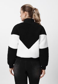 Mustvalge fliisist jakk lühendatud lõikega. Omadusteks on ševroonimuster, kõrge krae ning soonilised mansetid ja alumine äär.