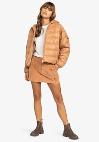Doudoune beige clair avec capuche, design matelassé et fermeture éclair, associée à une jupe en velours côtelé beige clair et des bottines marron.