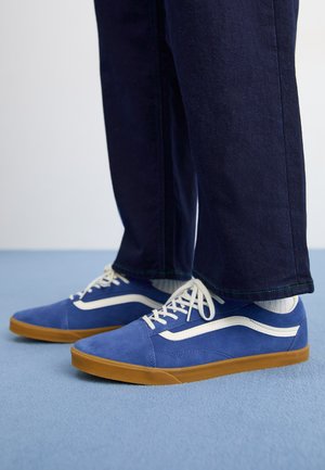Zapatillas de gamuza azul con acentos blancos y suela de goma. Combinadas con pantalones de mezclilla oscuros, mostrando el diseño y los detalles del material del zapato.