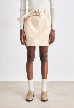 Beige minirok van nepleder met twee brede riemen en zilveren gespen. Glad oppervlak met naden aan de voorkant, gecombineerd met roomkleurige geribbelde sokken en beige loafers.