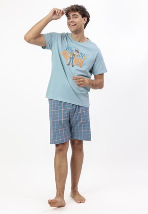 Joven sonriente con camiseta azul con la frase "¡Hola, compañero!" y pantalones cortos a cuadros azules, descalzo, posando con una mano levantada cerca de su cabeza.