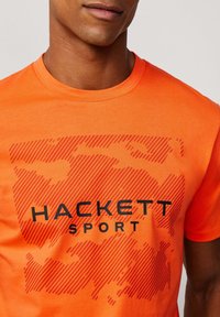 Oranžové bavlněné tričko s výrazným černým logem "HACKETT SPORT", s texturovaným designem zkřížených linií a vzorů na přední straně.