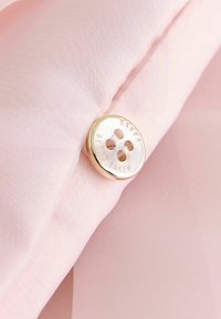 Tela rosa claro con una textura suave y un botón dorado brillante con texto grabado. El botón tiene cuatro orificios y forma circular.