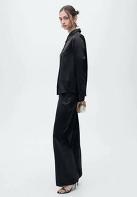 Cămașă elegantă din satin negru cu butoni, asortată cu pantaloni largi negri. Modelul ține în mână o poșetă transparentă și poartă pantofi cu toc subțire negri.