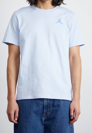 Person iført en lyseblå t-shirt med korte ærmer og et lille blåt Jumpman-logo på venstre side af brystet, kombineret med blå jeans.