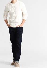 Homme portant un pull blanc à maille torsadée, un jean bleu foncé avec les revers relevés, et des chaussures en daim beige, debout les mains dans les poches.