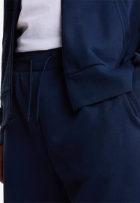 Ensemble sportif bleu marine comprenant une veste zippée et un pantalon à cordon de serrage, fabriqué en tissu doux et texturé avec des accents côtelés à la taille.