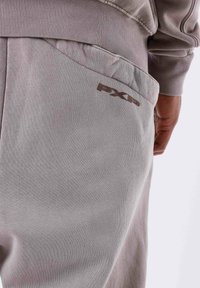 Grijze joggingsbroek met een zachte textuur, voorzien van een zijzak en een bruine "PXP"-logo op de achterkant. Elastische tailleband voor comfort.