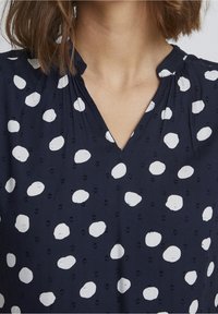 Navy blouse with a V-neckline, featuring white polka dots and subtle textured fabric. Gathered detailing at the neckline adds dimension.