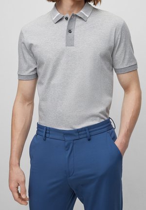 Man in een lichtgrijze getextureerde polo met donkergrijze afwerking en een blauwe op maat gemaakte broek, staande met één hand in zijn zak.