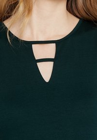 Tmavě zelený top s výstřihem ve tvaru scoop a detaily s výřezem. Měkká látka s hladkým povrchem a přiléhavý střih.