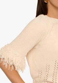 Cremfarbener Pullover mit strukturiertem Strick, der Fransendetails an den Ärmeln und Lochmustern am Saum aufweist.