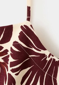 Gros plan d'un tissu beige avec un imprimé floral abstrait rouge foncé et une fine bretelle, montrant les détails des coutures du vêtement.