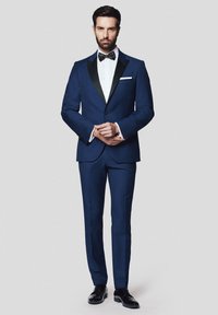 Granatowy garnitur z czarnymi klapami, biała koszula i czarny muszka. Dopasowany krój o gładkiej fakturze, noszony z wypolerowanymi czarnymi butami.