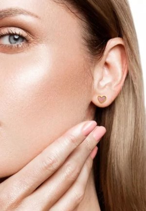 Gros plan sur le visage d'une femme à la peau claire, un œil bleu, la main touchant sa joue, portant une petite boucle d'oreille en forme de cœur en or.